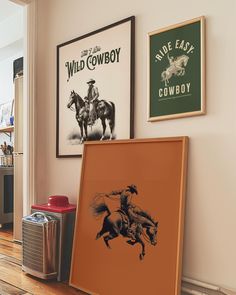 two framed pictures hang on the wall next to an old cowboy poster and other items