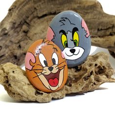 two rocks with cartoon cats painted on them sitting next to each other in front of driftwood