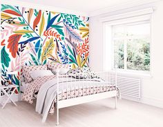 a white bed sitting under a window next to a wall covered in colorful flowers and leaves