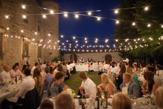 a large group of people sitting at tables in an outdoor area with string lights strung across the ceiling