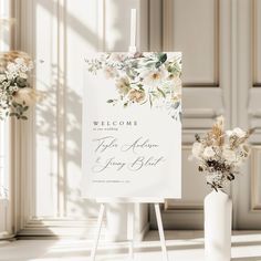 a welcome sign and vase with flowers in the background on an easel next to a white wall