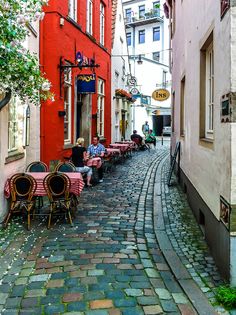 people sitting at tables in the middle of an alley with red buildings and cobblestone streets