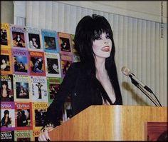 a woman standing at a podium with a microphone in front of her and posters on the wall behind her