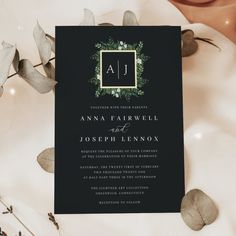 a black and green wedding card on top of a table with greenery around it