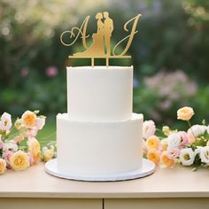 a white wedding cake with a gold monogrammed topper and flowers on the table