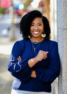 a woman leaning against a pole smiling at the camera with her arms crossed and wearing a blue top