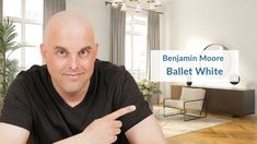 a bald man pointing at the camera in front of a living room with white walls and wood floors