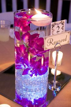 purple flowers are in a glass vase with water and lit candles on the table top