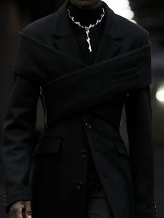 a man in a black coat and tie walking down the street with his hand on his hip