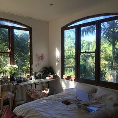 an unmade bed in a room with three windows and plants on the wall next to it