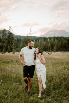 a man and woman standing in the grass with mountains in the background during their engagement session