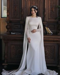 a woman wearing a white gown and cape standing in front of a wooden cabinet with books on it