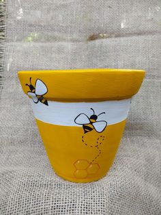 a yellow and white flower pot with bees painted on the side, sitting on a burlap surface