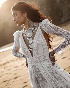 a woman standing on the beach wearing a white dress with intricate details and necklaces