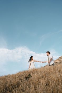 a man and woman holding hands while standing on top of a grass covered hill under a blue sky