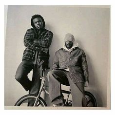 two men sitting on top of bikes in front of a wall with their arms crossed