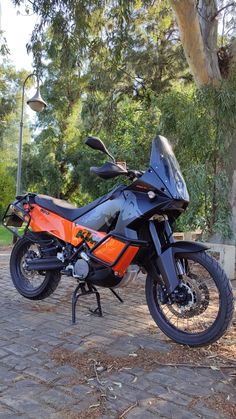 an orange and black motorcycle parked on the side of a brick road next to a tree