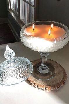 two candles are lit in a glass bowl on a wooden stand next to a window