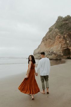 a man and woman holding hands walking on the beach with an ocean in the background