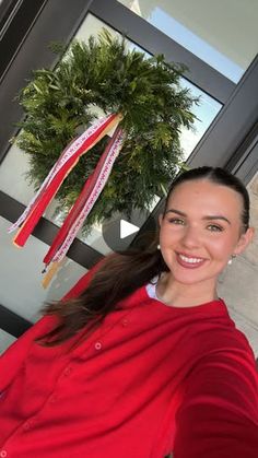 a woman in a red shirt is smiling and holding up a wreath with ribbons hanging from it