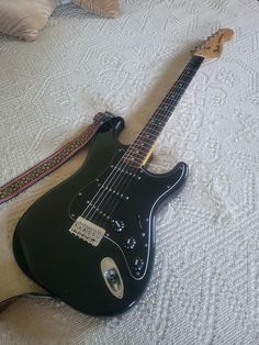 a black electric guitar laying on top of a white bedspread next to pillows