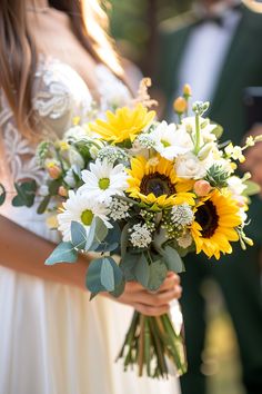 a bride holding a bouquet of sunflowers and greenery with her groom in the background