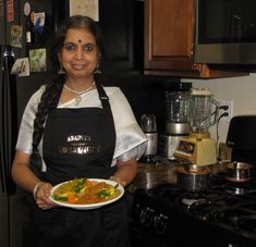 a woman in an apron holding a plate of food next to a stove top oven