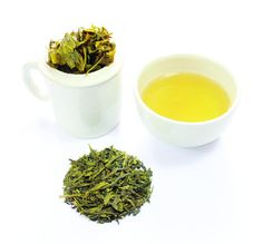 a white cup filled with green tea next to a pile of leaves and a bowl of yellow tea