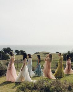 a group of women in long dresses walking across a lush green field next to the ocean