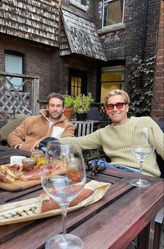 two men sitting at an outdoor table with food and wine glasses in front of them