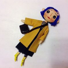 a doll with blue hair wearing a yellow coat and black dress, holding a purse