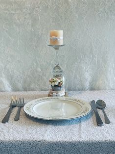 an empty plate and silverware on a white table cloth with a glass vase in the center