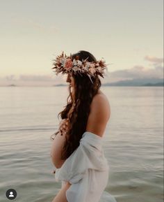 a pregnant woman wearing a flower crown standing in the water with her back to the camera