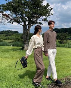 a man and woman holding hands while walking in the grass with a tree behind them