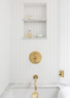 a bathroom with white tile and gold faucet, shelving above the bathtub