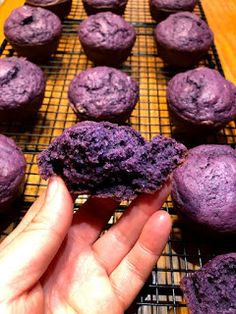 a person holding up a purple muffin in front of some other muffins on a cooling rack