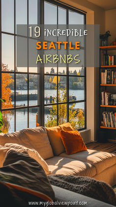 A cozy living room in a Seattle Airbnb with large floor-to-ceiling windows overlooking a serene waterfront view, framed by autumn trees and golden sunlight casting warm shadows across a plush sofa with colorful throw pillows.