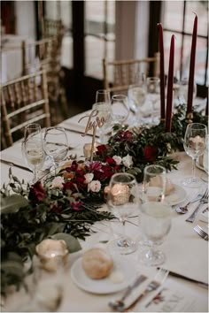 the table is set with white plates and silverware, red flowers, greenery and candles