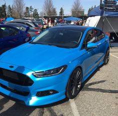 a blue car parked in a parking lot next to other cars with people standing around