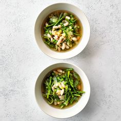 two white bowls filled with green vegetables and bean soup on top of a gray table