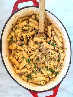 a red pot filled with pasta and meat on top of a white countertop next to a wooden spoon