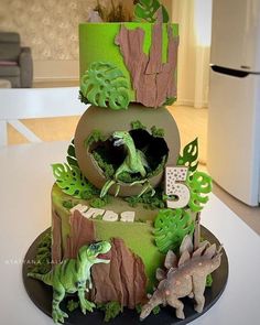 a three tiered cake decorated with green and brown icing, dinosaur figures and trees