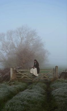 a woman sitting on top of a wooden fence next to a field covered in fog