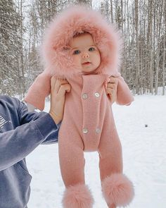 a woman holding up a baby wearing a pink snowsuit and fur hat in the snow