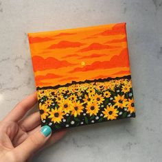 a hand holding an orange and black painted canvas with sunflowers in the background