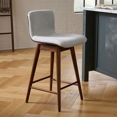 a wooden bar stool with a grey upholstered seat and back rests on a hard wood floor in front of a white brick wall