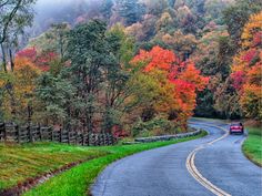 a red car driving down a road surrounded by fall colored trees in the mountains behind it