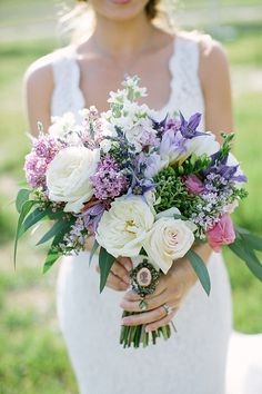 a woman holding a bouquet of white and purple flowers on her wedding day in the grass