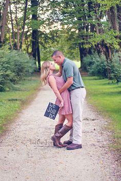 a man and woman kissing on the side of a dirt road with trees in the background