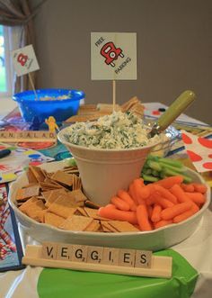 a bowl of dip surrounded by crackers and veggies on a table with other snacks